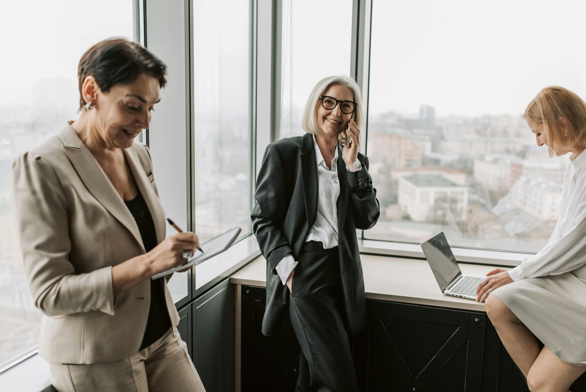 Trois femmes en costume discutent près d'une grande fenêtre avec vue sur la ville.